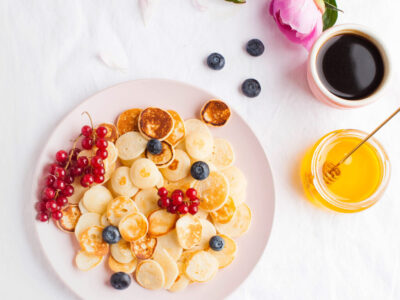 Tiny pancakes with berries, honey and flowers on a white tablecloth and space for text. Pancake cereal. The concept of Breakfast, food trends. Copy space. . High quality photo