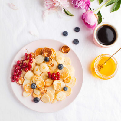 Tiny pancakes with berries, honey and flowers on a white tablecloth and space for text. Pancake cereal. The concept of Breakfast, food trends. Copy space. . High quality photo